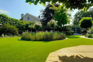 Een prachtige tuin die is aangelegd met veel ronde vormen en beplanting waar er kunstgras is aangelegd door het kunstgrascentrum. In het midden heb je een cirkel met beplanting.