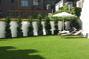 Een dakterras aangelegd met kunstgras door het kunstgrascentrum waar er een mevrouw aan het genieten is op de ligzetel op kunstgras met een prachtige zon. Er staat ook veel beplanting op het kunstgras en een standbeeld van een wit konijn.