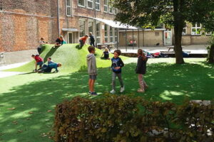 Kinderen die aan het spelen zijn op de speelplaats aangelegd met kunstgras door het kunstgrascentrum. Er zijn ook bomen verwerkt in het kunstgras.