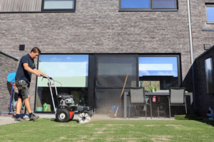 Een werkman van het kunstgrascentrum die bezig is met het borstelen van het kunstgras in een achtertuin met op de achtergrond een terras waar er een tuinset op staat.