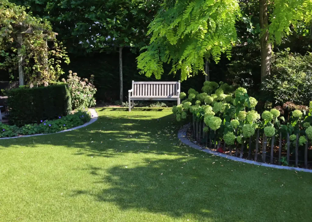 Foto van een kunstgras siertuin met veel beplanting rondom een zithoek met houten tuinbank